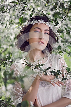 portrait of a young beautiful woman in white clothes standing next to a blooming cherry tree in spring. a wreath on the