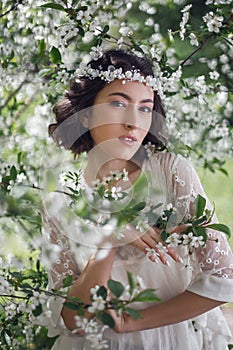 portrait of a young beautiful woman in white clothes standing next to a blooming cherry tree in spring. a wreath on the