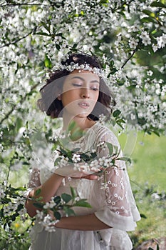 portrait of a young beautiful woman in white clothes standing next to a blooming cherry tree in spring. a wreath on the