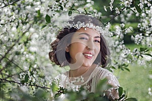 portrait of a young beautiful woman in white clothes standing next to a blooming cherry tree in spring. a wreath on the