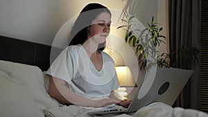 Portrait of young beautiful woman is working at laptop lying in bed. Cozy home atmosphere in evening. Concept of remote