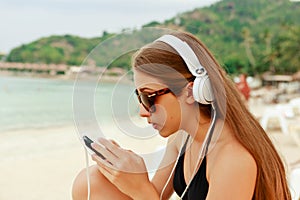 Portrait of a young beauty teenager girl sitting by white sand beach listening to music using headphones and smartphone against on