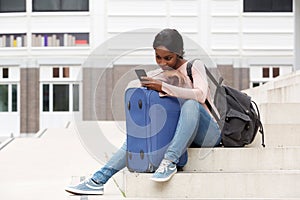 Young black woman sitting on steps with suitcase bag and looking at mobile phone