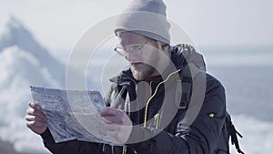 Portrait of young blond bearded handsome man in warm jacket and hat standing on the glacier checking with the map