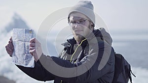 Portrait of young blond bearded handsome man wearing warm jacket and hat standing on the glacier checking with the map