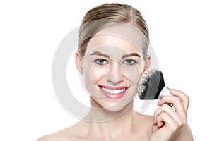 Portrait of a young blond woman applying dry cosmetic tonal foundation on her face using make up brush. Beauty portrait.