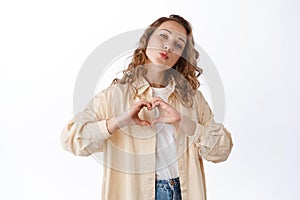 Portrait of young blond woman with curly hair, showing love heart gesture, pucker lips, say I love you, express