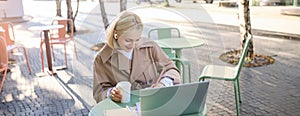 Portrait of young blond woman, female student in street cafe, wearing wireless headphones, using laptop, having online