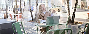 Portrait of young blond woman, female student in street cafe, wearing wireless headphones, using laptop, having online