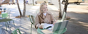 Portrait of young blond woman, female student in street cafe, wearing wireless headphones, using laptop, having online