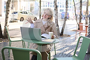 Portrait of young blond woman, female student in street cafe, wearing wireless headphones, using laptop, having online