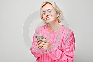 Portrait of young blond woman in pink sweater holding mobile phone in hand with happy smiling face. Person with smartphone
