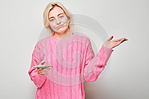 Portrait of young blond woman in pink sweater looks on mobile phone and thinks. Person with smartphone