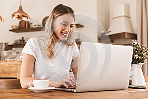 Portrait of young blond woman working on laptop and drinking coffee while sitting in cozy cafe indoor