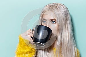 Portrait of young blonde girl with blue eyes drinking tea from black cup of background of aqua menthe color.