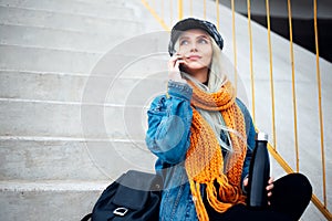 Portrait of young blonde girl, looking up, sitting on the urban stairs, talking on smartphone, holding reusable, steel thermo wate