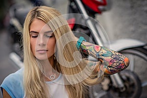 Portrait of a young blonde girl sitting on the background of the motorcycle. Women`s hands with a tattoo. Urban fashion.