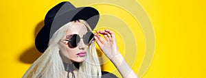 Portrait of young blonde girl wearing black choker, hat and sunglasses on yellow background.