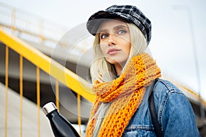 Portrait of young blonde teenager girl, wearing blue denim jacket, with orange scarf and plaid cap, holding a black steel bottle.