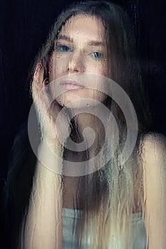 Portrait of young blonde woman behind the window glass with raindrops. S