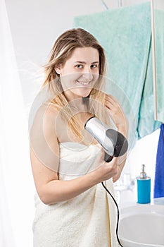 Portrait of young blonde woman drying hair with hairddryer and looking in camera