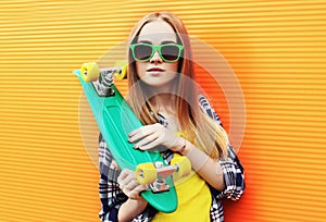 Portrait young blonde woman with green skateboard wearing a sunglasses over colorful orange wall