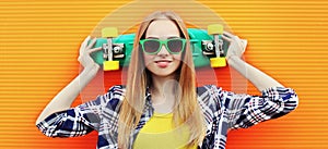 Portrait young blonde woman with green skateboard wearing a sunglasses over colorful orange wall