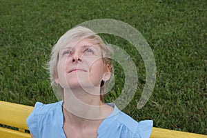 Portrait of a young blonde woman with a happy smiling face looking up. Female in a blue dress sitting on the bench