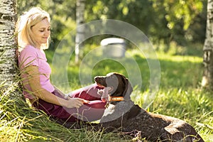 Portrait of a young blonde woman sitting, relaxing on the grass near a tree in the park with a hunting dog of the Kurz-Haar breed