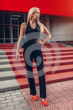 Portrait of young blonde woman wearing stylish clothing and accessories by modern business center in city.