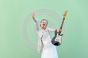 Portrait of young blonde woman in white shirt, skirt, and striped blouse and electric guitar standing with rock sign tongue out