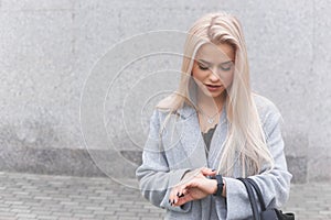 Portrait of young blondhair elegantly dressed woman in a coat uses a smart bracelet standing outdoors