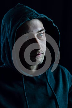 Portrait of a young blue-eyed man with a hood covering his head who in the shadow is staring away