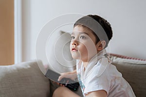 Portrait young boy holding remote control and looking up with curious face, Kid sitting on sofa watching cartoon on TV, Child