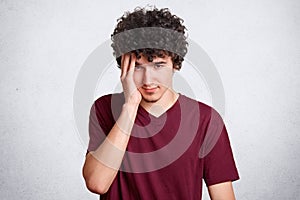 Portrait of young boy with little beard and curly dark hair, keeps hand on temple  on white wall, sufers from headache,