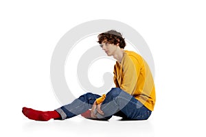 Portrait of young boy sitting on floor in sadness, feeling asocial over white background. Loneliness, depression