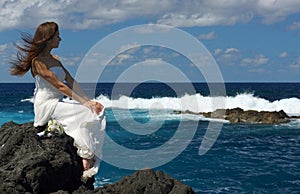 Portrait of young bride in wedding dress sitting on ocean rock shore and looking at ocean. Wedding ceremony on ocean