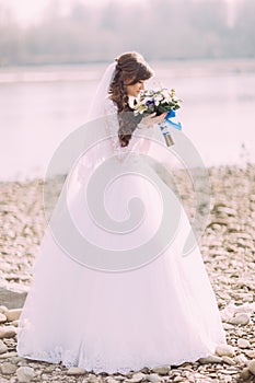 Portrait of young bride in white dress and veil sniffing wedding bouquet with blue bow outdoors