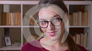 Portrait of young brunette female student with ponytail in eye glasses smiles with flirtation into camera in library.