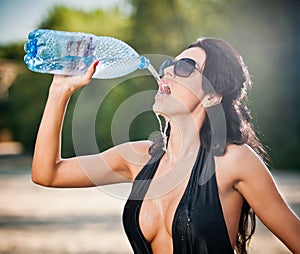 Portrait of young brunette girl in black low-cut swimsuit drinking water from a bottle. Sensual attractive woman