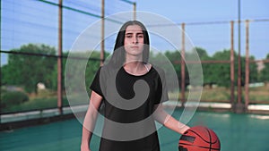 Portrait young brunette girl holding a basketball ball looking at the camera standing on the basketball court outdoors.