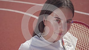 Portrait young brunette girl sits at outdoor basketball court holding a tennis racket in sunny summer weather. Concept