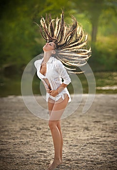 Portrait of young brunette girl in white swimsuit and male shirt posing on a beach with a forest in background