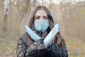 Portrait of young brunette woman in blue protective mask and rubber gloves shows stop gesture outdoors in spring wood