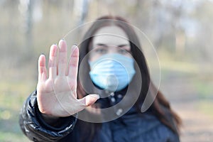 Portrait of young brunette woman in blue protective mask shows stop gesture outdoors in spring wood