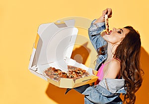 Portrait of young brunette woman in pink top and denim jacket holding a box of fresh tasty pizza eating a slice