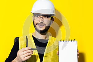 Portrait of young builder engineer with safety helmet and glasses, explains showing a pen on notebook with mockup on yellow.