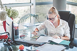 Portrait of young business lady in glasses sitting at her workplace and reads documents. Office worker at modern office workplace