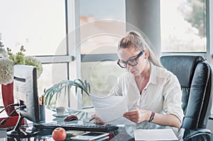 Portrait of young business lady in glasses sitting at her workplace and reads documents. Office worker at modern office workplace