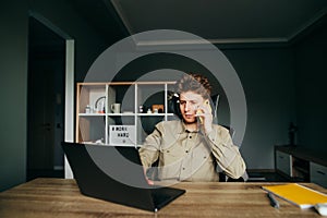 Portrait of a young business man working on laptop at home, using the Internet and talking on the phone with a serious face on the